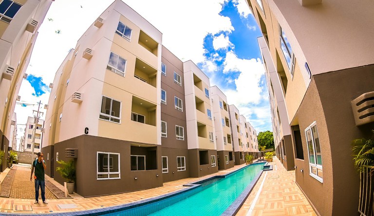 P´rédio marrom e bege e uma piscina na cor azul. Na imagem há um homem vestindo uma calça jeans e blusa azul.