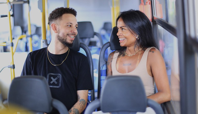 Na foto casal conversa alegremente sentados nos bancos de um ônibus.