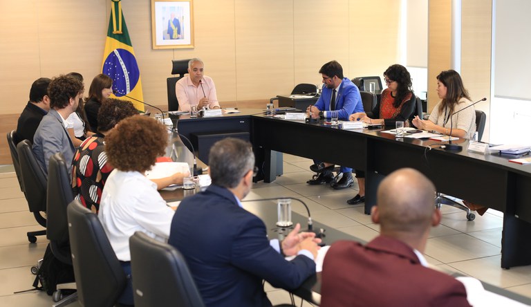 Grupo de pessoas sentados reunidos numa mesa redonda. Ao fundo um quadro com a foto de um homem branco com uma faixa verde e amarela.
