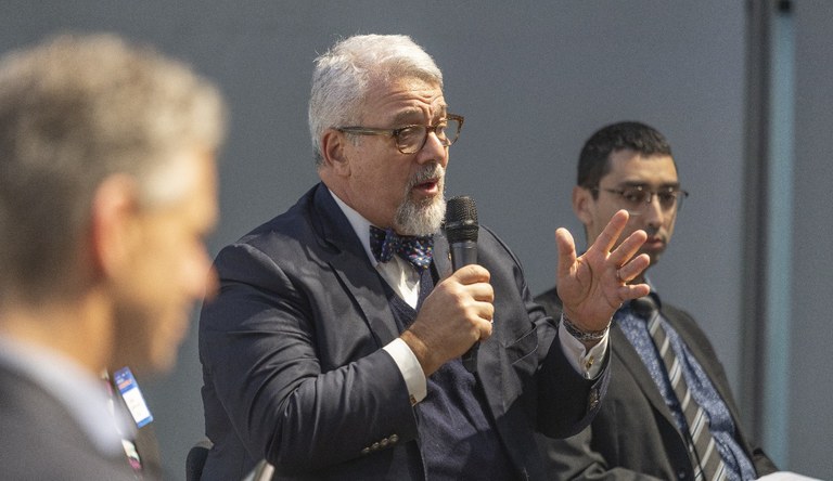 Na foto dois homens observam um terceiro falando ao público