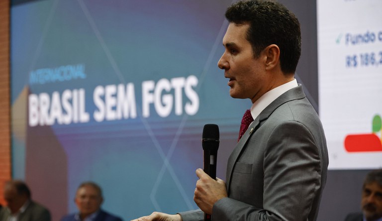 Homem branco, alto com terno cinza. Ao fundo um painel azul com a frase: Brasil sem FTGS.