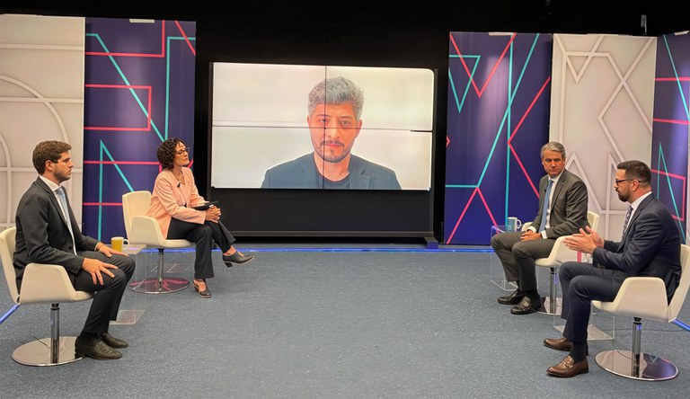 Na foto três homens e uma mulher debatem junto a outro em um telão.