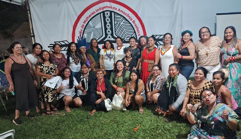 Grupo de mulheres indígenas e negras, algumas estão em pé e outras sentadas numa grama, Ao fundo, um banner com a frase: mulheres in´digenas.