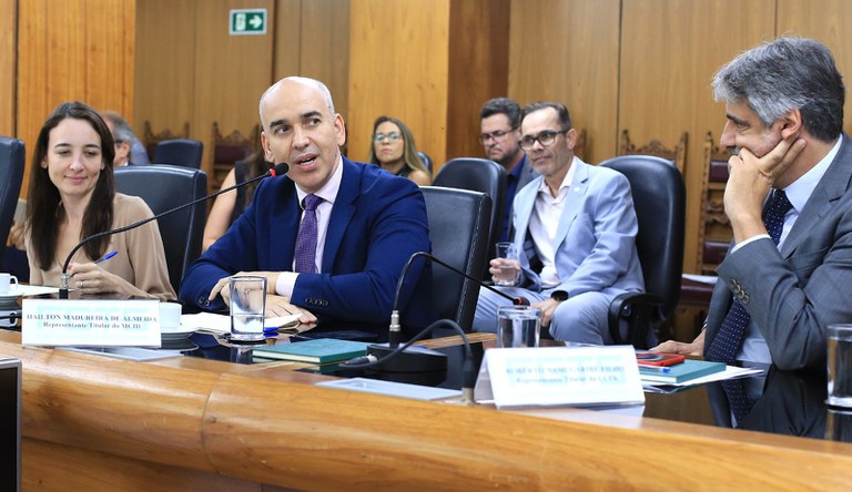 Dois homens, um deles é careca. Os dois usam blazer nas cores azul e cinza. Ao lado esquerdo uma mulher branca com cabelos grande veste blusa branca.