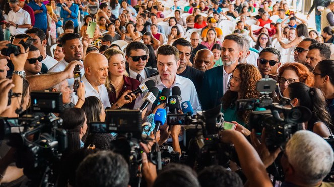 Um homem alto, de pele branca, veste uma blusa social azul e está posicionado em frente a vários microfones. Ao seu redor, há jornalistas com câmeras, gravadores e blocos de anotações, demonstrando que ele está concedendo uma entrevista coletiva. 