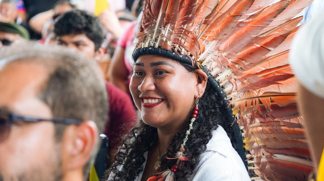 Mulher indígena com cocar vermelho sorrindo. 