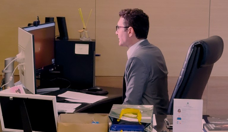 Homem branco sentado em frente a dois computadores participa de reunião online. Veste um blazer cinza e usa óculos de grau.