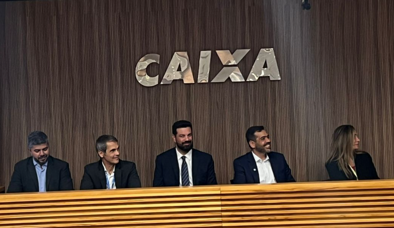 Quatro homens e uma  mulher sentados numa bancada, ao fundo o logo da Caixa Econômica Federal.