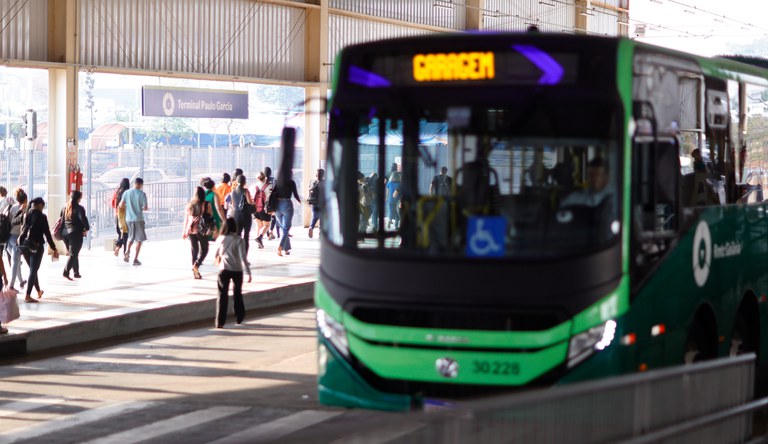 Na imagem pessoas transitando numa rodoviária e um ônibus verde parado.