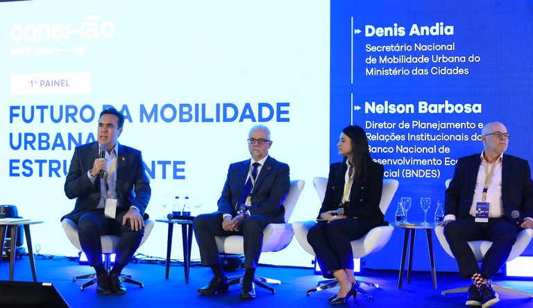 Na imagem quatro pessoas sentadas,, uma delas com microfone na mão