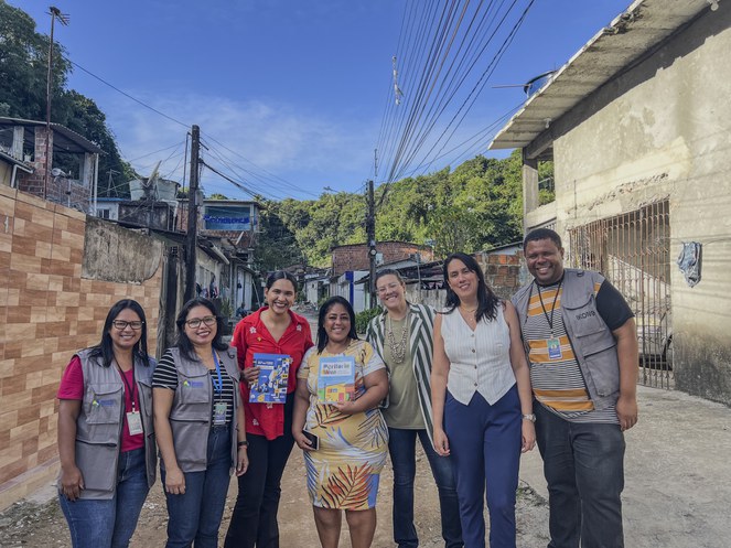 Na imagem, um grupo de pessoas sorri para a câmera. Ao fundo, aparecem casas da comunidade em Pernambuco