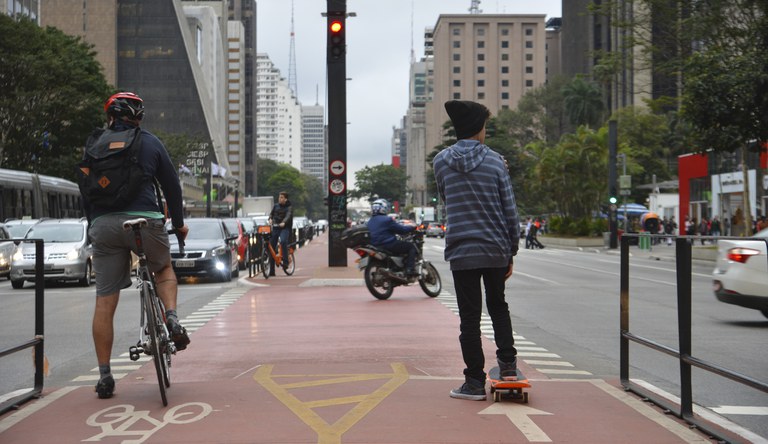 Na foto pessoas de bicicleta e skate aguardando o sinal fechar para atravessar a avenida