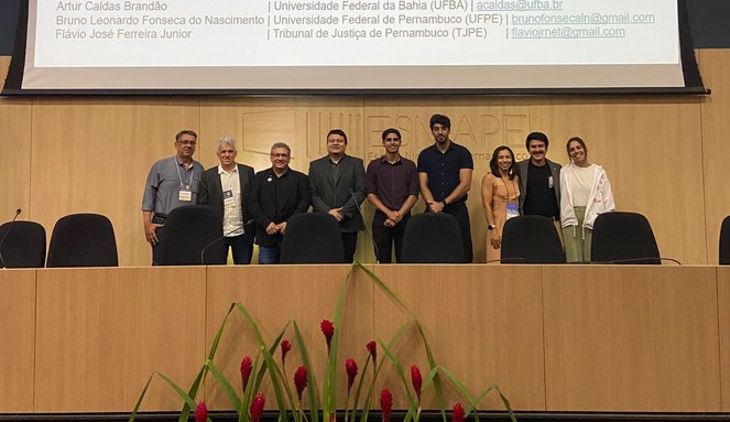 Pessoas em pé sorrindo para fotógrafo, ao fundo telão com nomes de participantes do evento 
