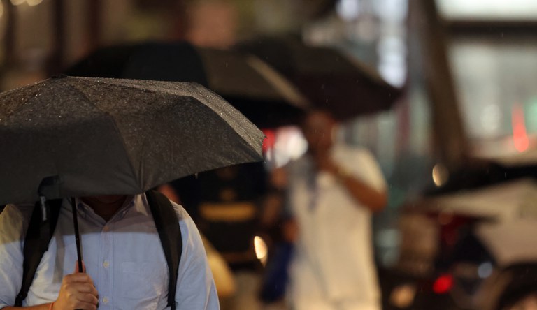 Na imagem pessoas andando na rua com guarda-chuva