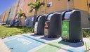 Na foto três lixeiras ecológicas com prédios do MCMV ao fundo