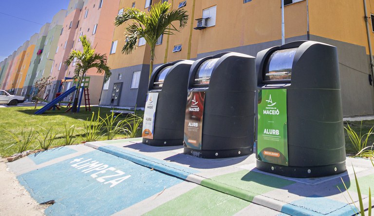 Na foto três lixeiras ecológicas com prédios do MCMV ao fundo
