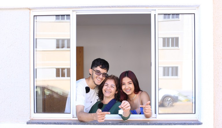 Na imagem mulher ao lado de dois jovens na janela de casa segurando a chave da casa p´rópria