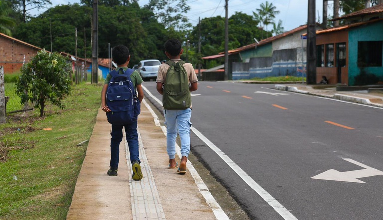 Duas crianças caminham pela calçada de via pavimentada