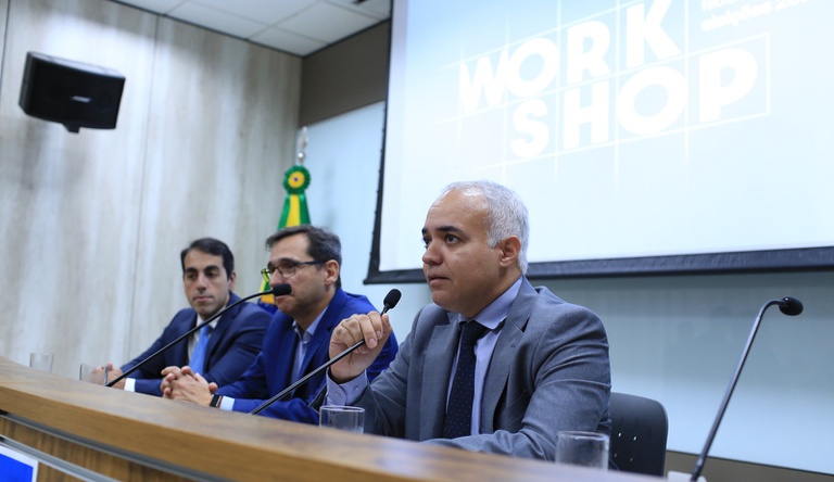 Na imagem dois homens sentados, um deles fala ao microfone.