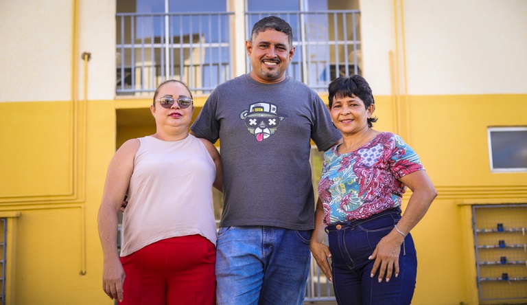 Na imagem um homem no centro e duas mulheres nas laterais posam para foto em frente a casa de alvenaria.