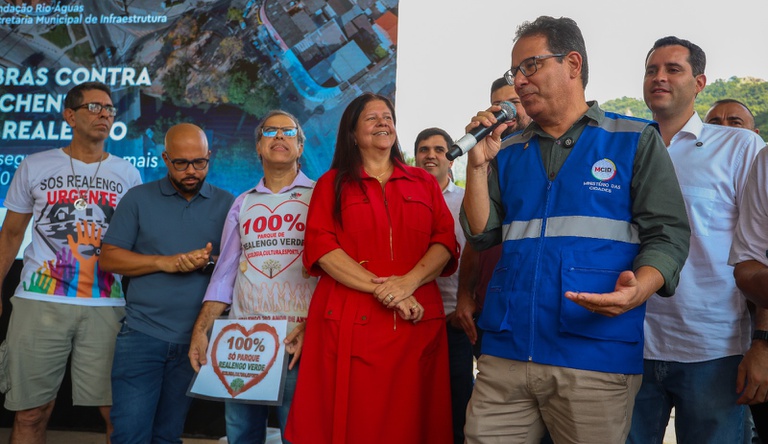 Na imagem homem segura microfone, ao lado pessoas sorriem.