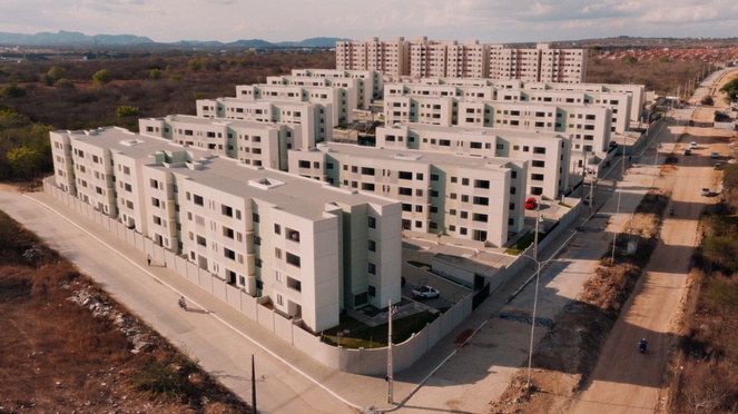 Na imagem foto panorâmica de prédio residencial, ao lado rua sem asfalto