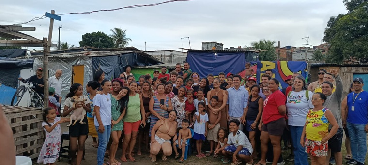 Na imagem grupo de pessoas posam para foto, ao fundo casa de madeira