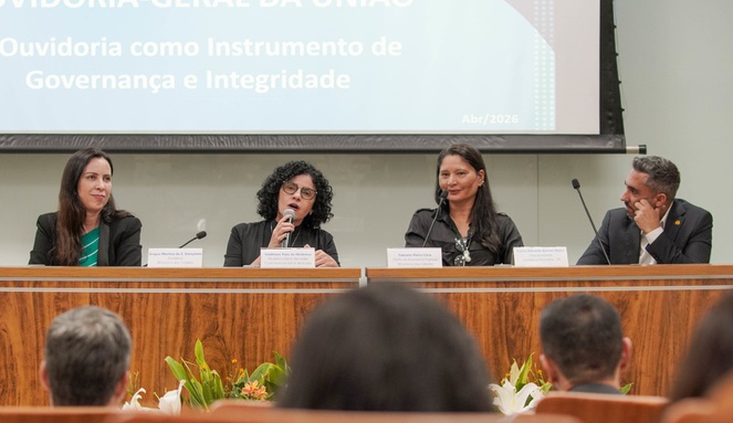 Na imagem três mulheres e um homem sentados numa bancada, uma delas fala ao microfone.