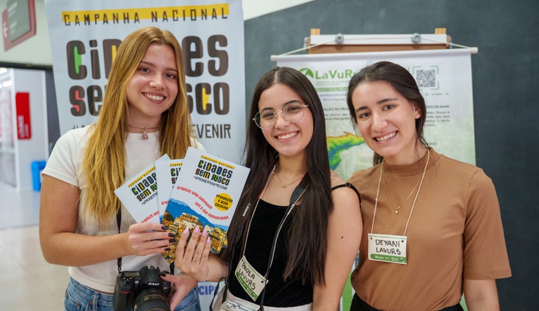 Na imagem três mulheres posam para foto segurando panfleto da campanha Cidades sem Risco