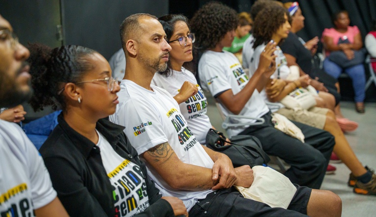 Na imagem grupos de pessoas sentadas ouvindo palestrante, todos vestem blusa da campanha Cidades sem Risco