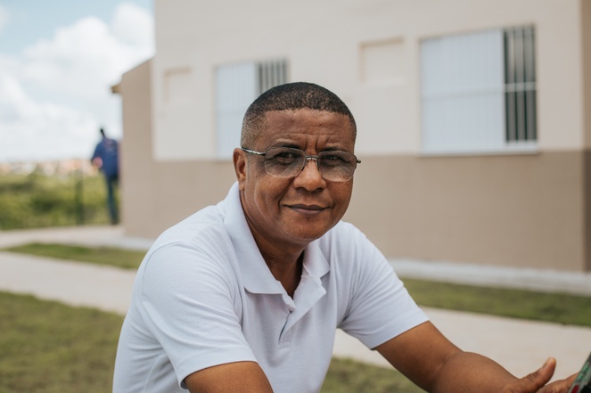 Na imagem homem sentado posa para foto em frente a sua nova casa