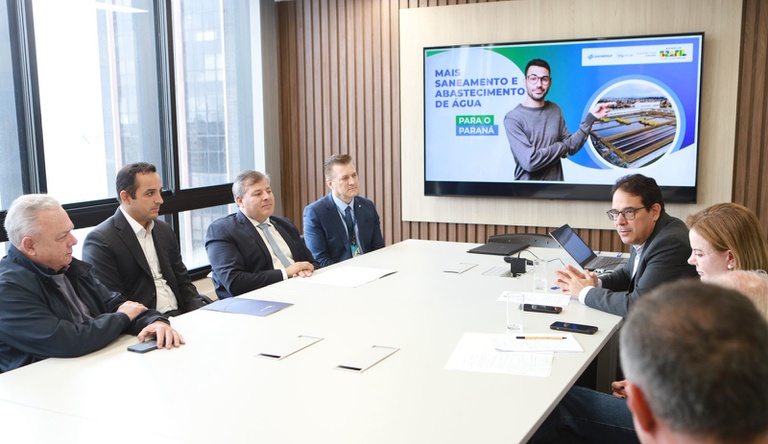 Na imagem pessoas sentadas durante reunião, ao fundo painel com dados do saneamento no Paraná