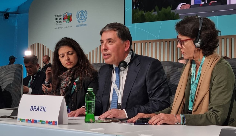 Na imagem homem ao lado de duas mulheres, ele discursa em evento sobre cidades resilientes