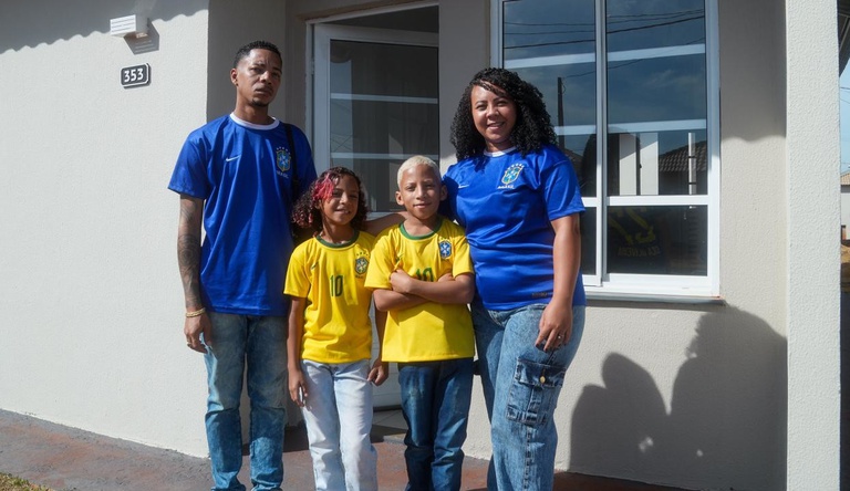 Na imagem família vestida com a blusa da seleção brasileira posa em frente a sua casa p´ropria.