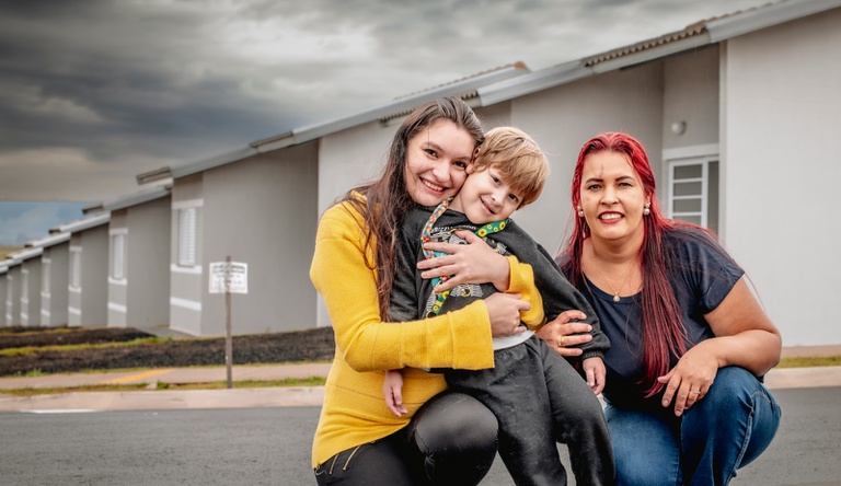 Na imagem duas mulheres abraçando uma criança comemorando a casa nova