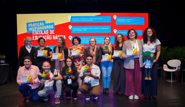 Na imagem vencedores do prêmio AprenderParaPrevenir posam para foto com prêmio e bandeiras dos estados.