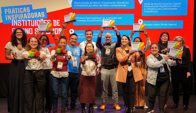 Na imagem pessoas posam para foto segurando prêmio da campanha AprenderParaPrevenir, ao fundo painel da campanha.