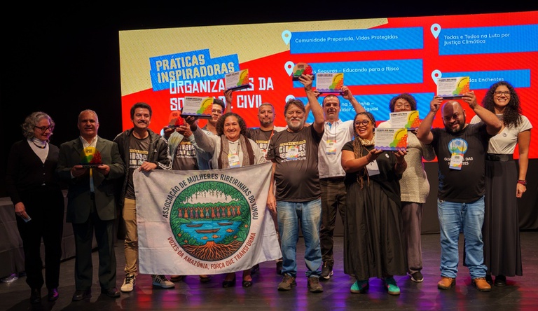 Na imagem mulheres posam segurando prêmio da campanha AprenderParaPrevenir, ao fundo painel de boas p´raticas