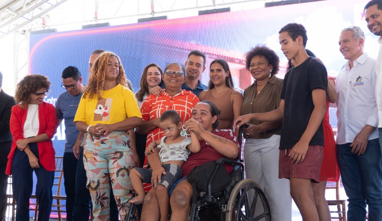 Na imagem mulher em cadeira de rodas segura criança no colo, ela segura chave da casa própria, ao lado pessoas sorrindo.