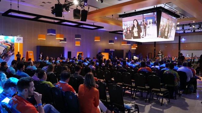 Na foto panoramica do local do evento com telões no teto e o público assistindo