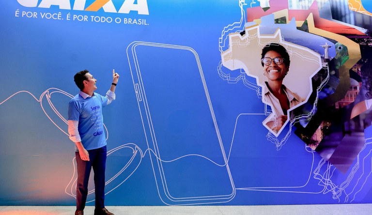 Na foto o ministro das cidades a frente de um telão azul da Caixa aponta para o alto na direção do nome Caixa