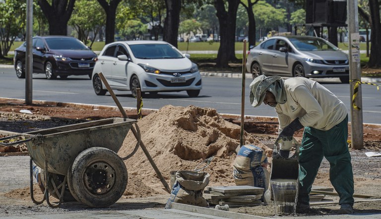 Na foto trabalhador da construção civil realiza revitalização de calçada a margem de uma via com carros trafegando.