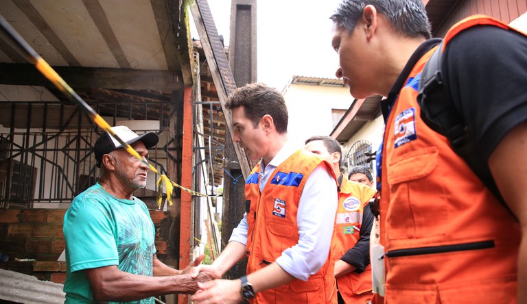 Três homens, dois vestindo colete laranja da Defesa Civil, o outro com boné e blusa verde.