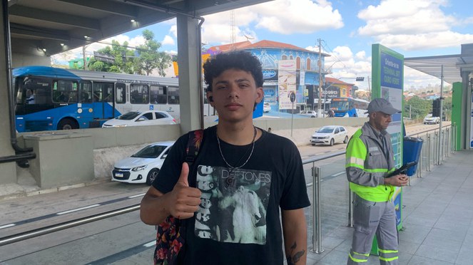 Menino sorrindo com polegar estendido em frente a uma estação BRT.