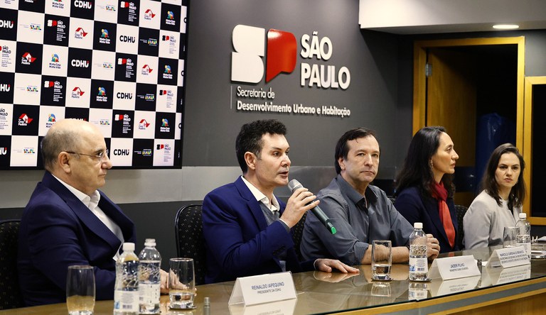 Na imagem três homens e duas mulheres sentados numa mesa, um deles segura o microfone para falar com o público.