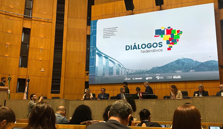 Na imagem há pessoas sentadas numa bancada palestrando para um grupo de pessoas sentadas.