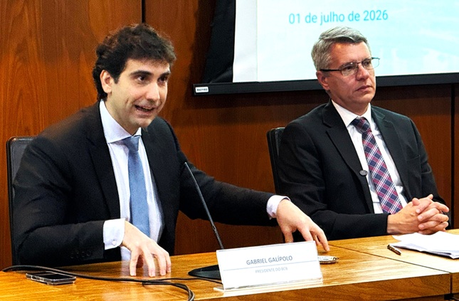 Dois homens de terno falam ao microfone em uma mesa de auditório.
