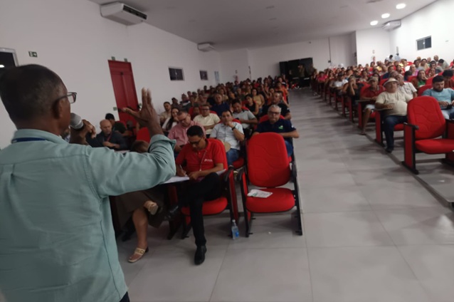 Palestrante fala ao público em auditório com cadeiras vermelhas.