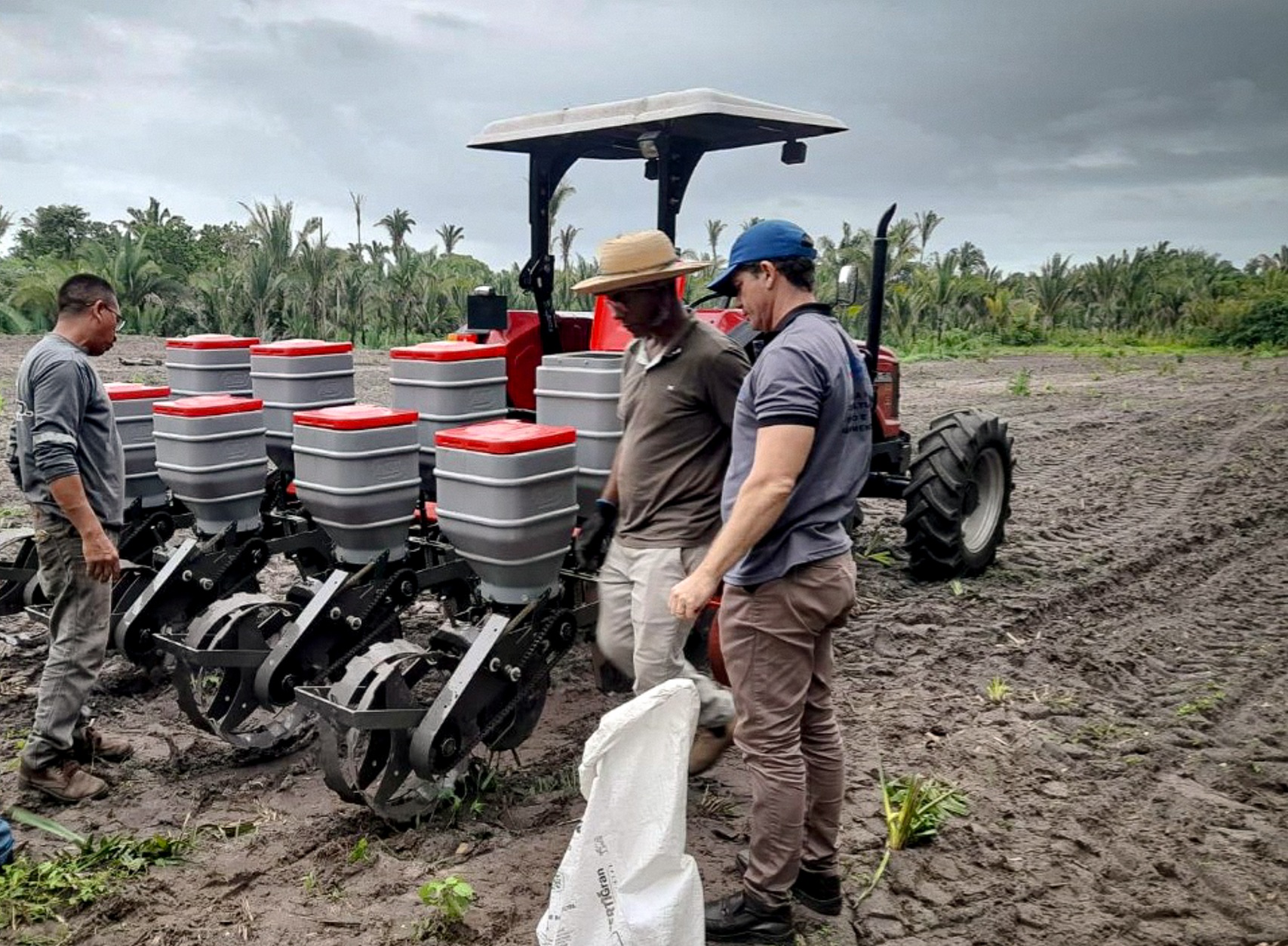 Três homens observam equipamento de plantio acoplado a um trator vermelho em área de terra arada.