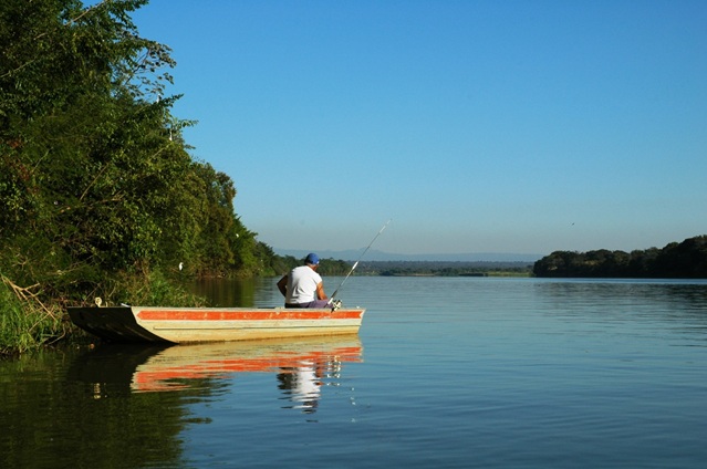 Homem em canoa pescando próximo à margem de rio cercada por vegetação